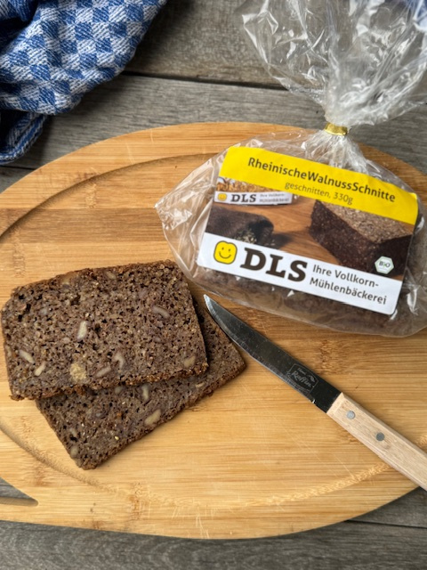 KI generiert: Das Bild zeigt ein rundes Stück Käse und zwei Scheiben Brot auf einem Holzbrett, begleitet von einem Messer. Im Hintergrund ist ein blau-weiß kariertes Tuch sichtbar.