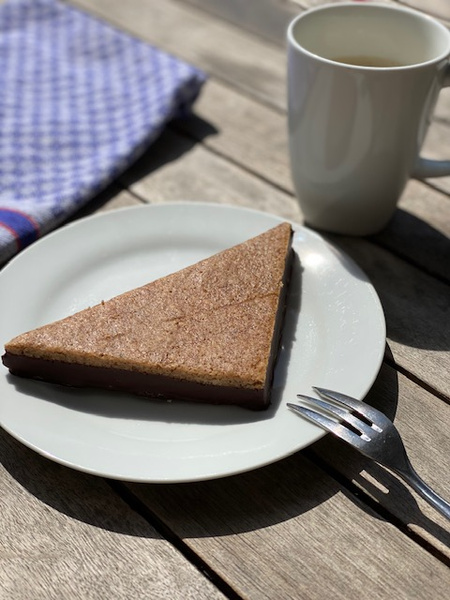 KI generiert: Das Bild zeigt ein rundes Stück Käse und zwei Scheiben Brot auf einem Holzbrett, begleitet von einem Messer. Im Hintergrund ist ein blau-weiß kariertes Tuch sichtbar.