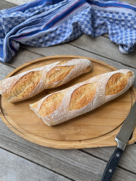 KI generiert: Zwei frische Baguettes auf einem Holzbrett mit Brotmesser, daneben ein blaues Geschirrtuch.