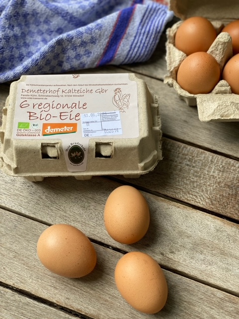 KI generiert: Das Bild zeigt ein rundes Stück Käse und zwei Scheiben Brot auf einem Holzbrett, begleitet von einem Messer. Im Hintergrund ist ein blau-weiß kariertes Tuch sichtbar.