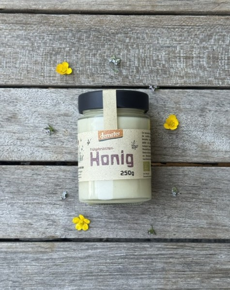 KI generiert: Ein Honigglas auf Holztisch, umgeben von kleinen gelben Blüten. Text: "Frühjahrstrachten Honig 250g".
