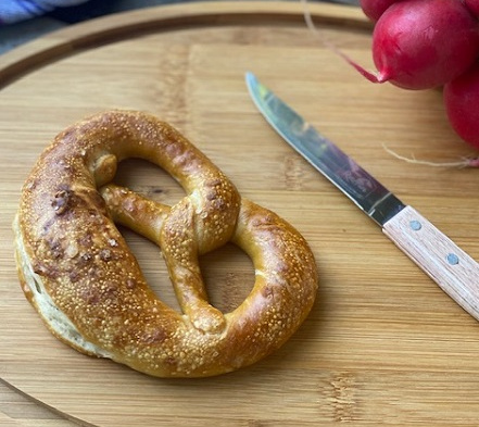 KI generiert: Das Bild zeigt ein frisch gebackenes, rundes Sauerteigbrot auf einem Holzbrett neben einem Brotmesser. Im Hintergrund ist ein blau-weiß kariertes Tuch zu sehen.