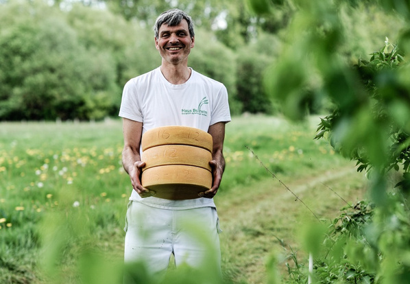 KI generiert: Ein Mann hält drei Käselaibe auf einer Wiese. Text: "Haus Bollheim".