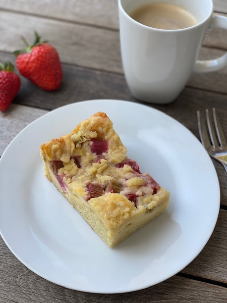 KI generiert: Ein Stück Kuchen auf einem Teller mit einer Tasse Kaffee und Erdbeeren im Hintergrund.