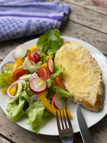 KI generiert: Das Bild zeigt ein frisch gebackenes Brotlaib auf einem Holzbrett, neben einem Brotmesser. Im Hintergrund ist ein blau-weiß kariertes Tuch zu sehen.