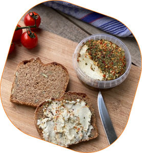 KI generiert: Auf dem Bild liegt ein Holzbrett mit zwei Scheiben Vollkornbrot, von denen eine mit einem Kräuteraufstrich bestrichen ist. Daneben befindet sich ein offener Becher mit dem Aufstrich und einige Tomaten.