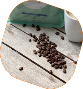 KI generiert: Das Bild zeigt verstreute Kaffeebohnen auf einem Holztisch neben einer Tasse Kaffee und einer Verpackung. Der Fokus liegt auf den Kaffeebohnen und der gemütlichen Kaffeeatmosphäre.